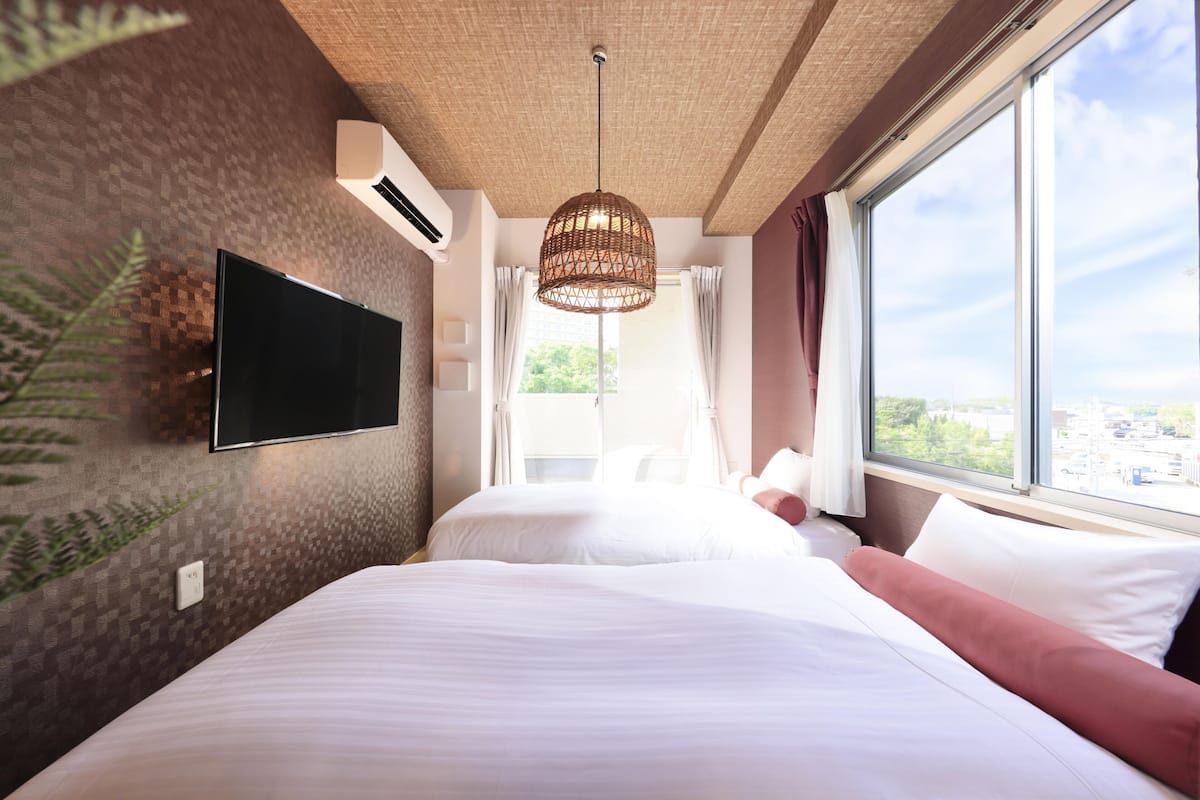 Une chambre d’hôtel avec un grand lit, un téléviseur fixé au mur, un ventilateur de plafond et une fenêtre donnant sur la ville.