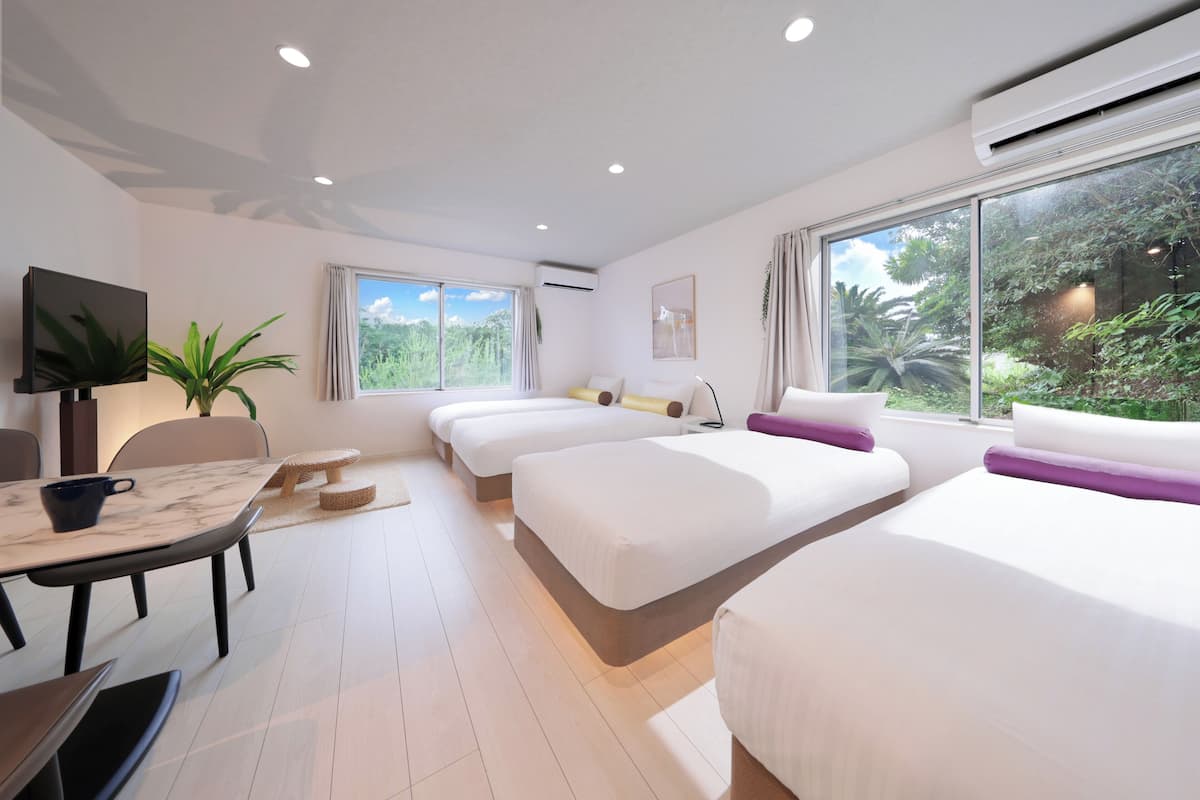 Une chambre d’hôtel moderne avec trois lits, un coin repas et de grandes fenêtres donnant sur un paysage verdoyant.
