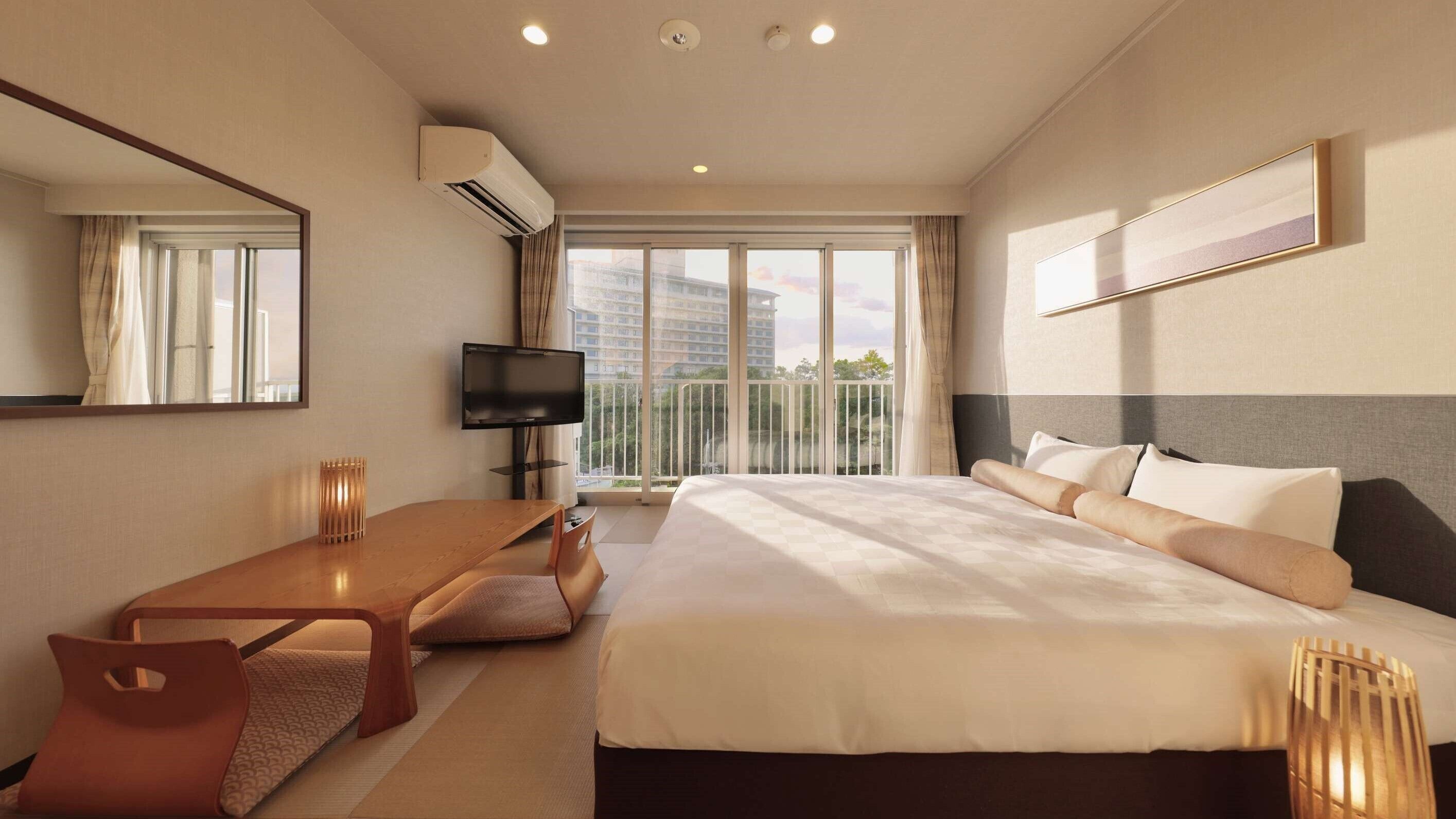 A hotel room with a bed, a desk, a TV, and a balcony with a view of buildings.
