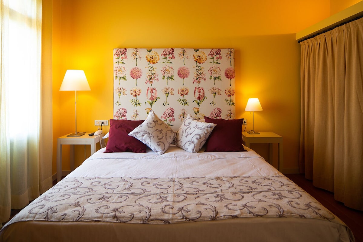 Een hotelkamer met een bed, twee bedlampen, een hoofdeinde met bloemenprint en een gordijn.