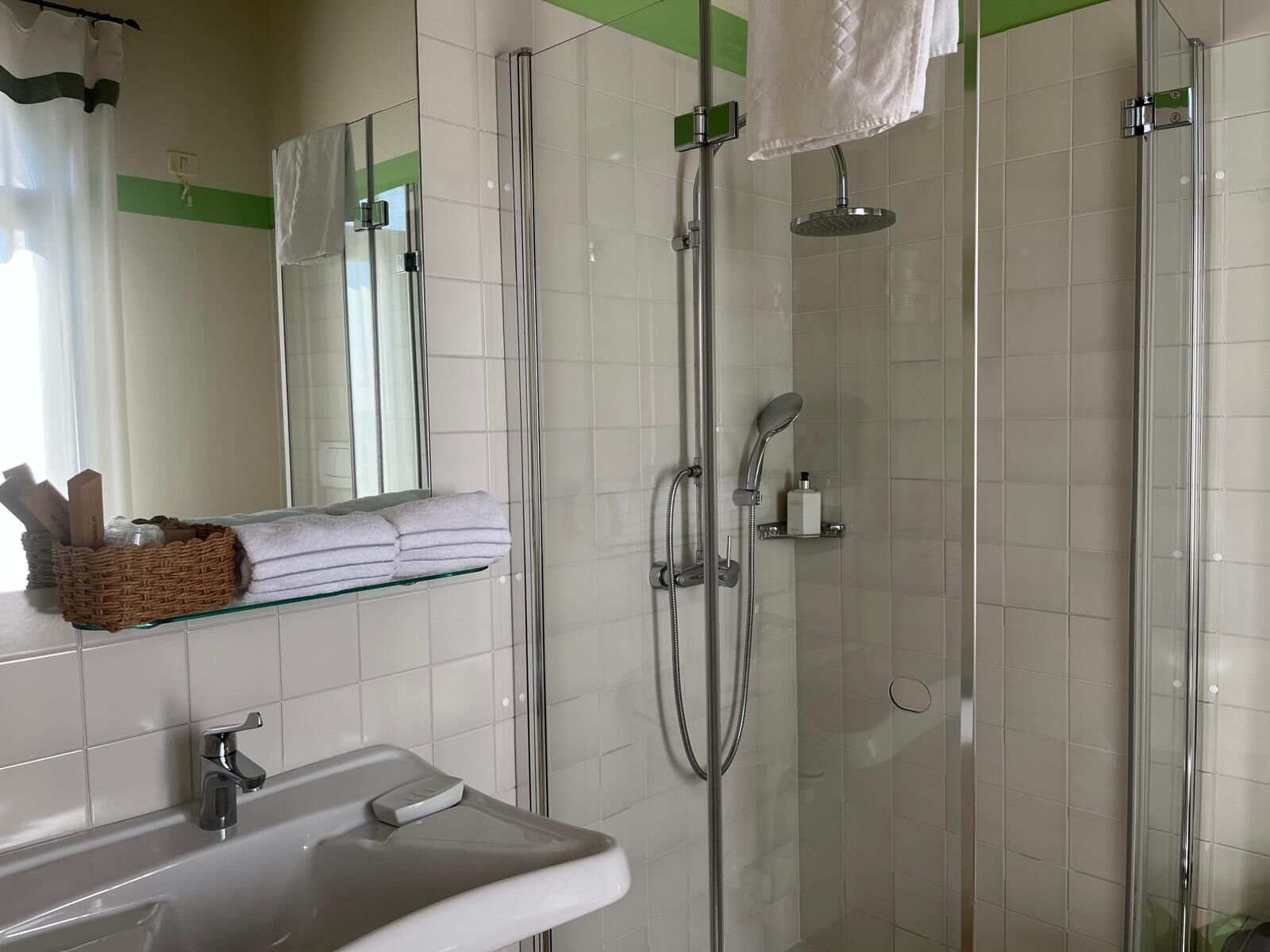 Un baño moderno con ducha envuelta en vidrio, repisa de vidrio con toallas y lavamanos con dispensador de jabón.