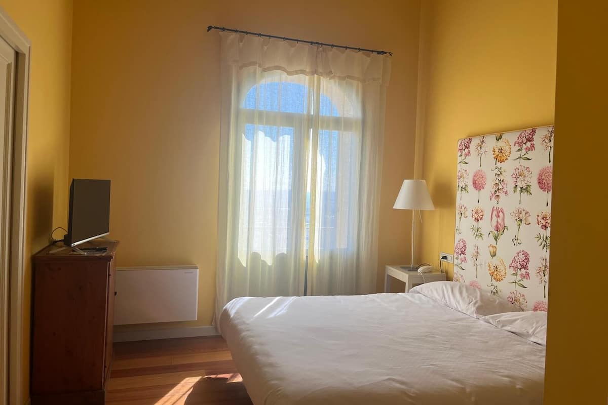 Een slaapkamer met een bed, een nachtkastje met een lamp, een radiator en een raam met gordijnen.