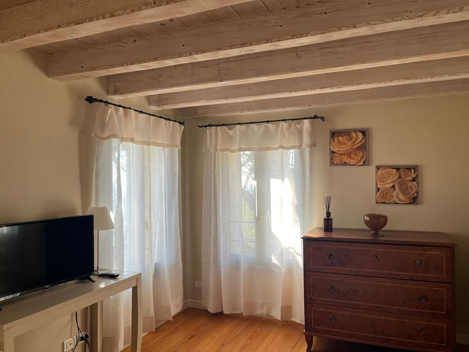 Ein Zimmer mit Holzbalken an der Decke, einem Fernseher auf einem Standfuß, einem hölzernen Schminktisch und transparenten Vorhängen.