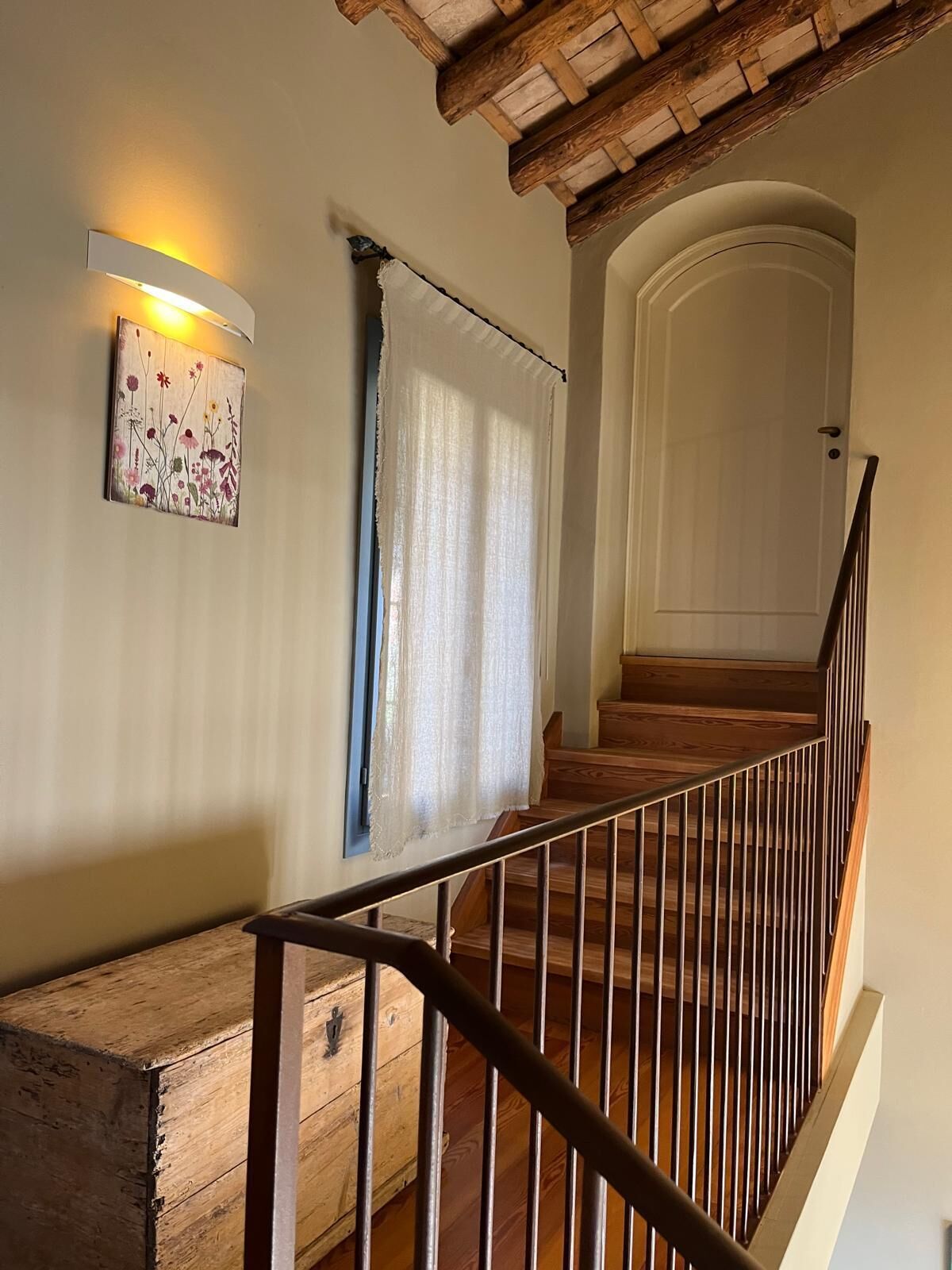 Una escalera de madera con barandilla de metal que lleva a una puerta blanca. Un cofre de madera está colocado en el suelo a la izquierda.