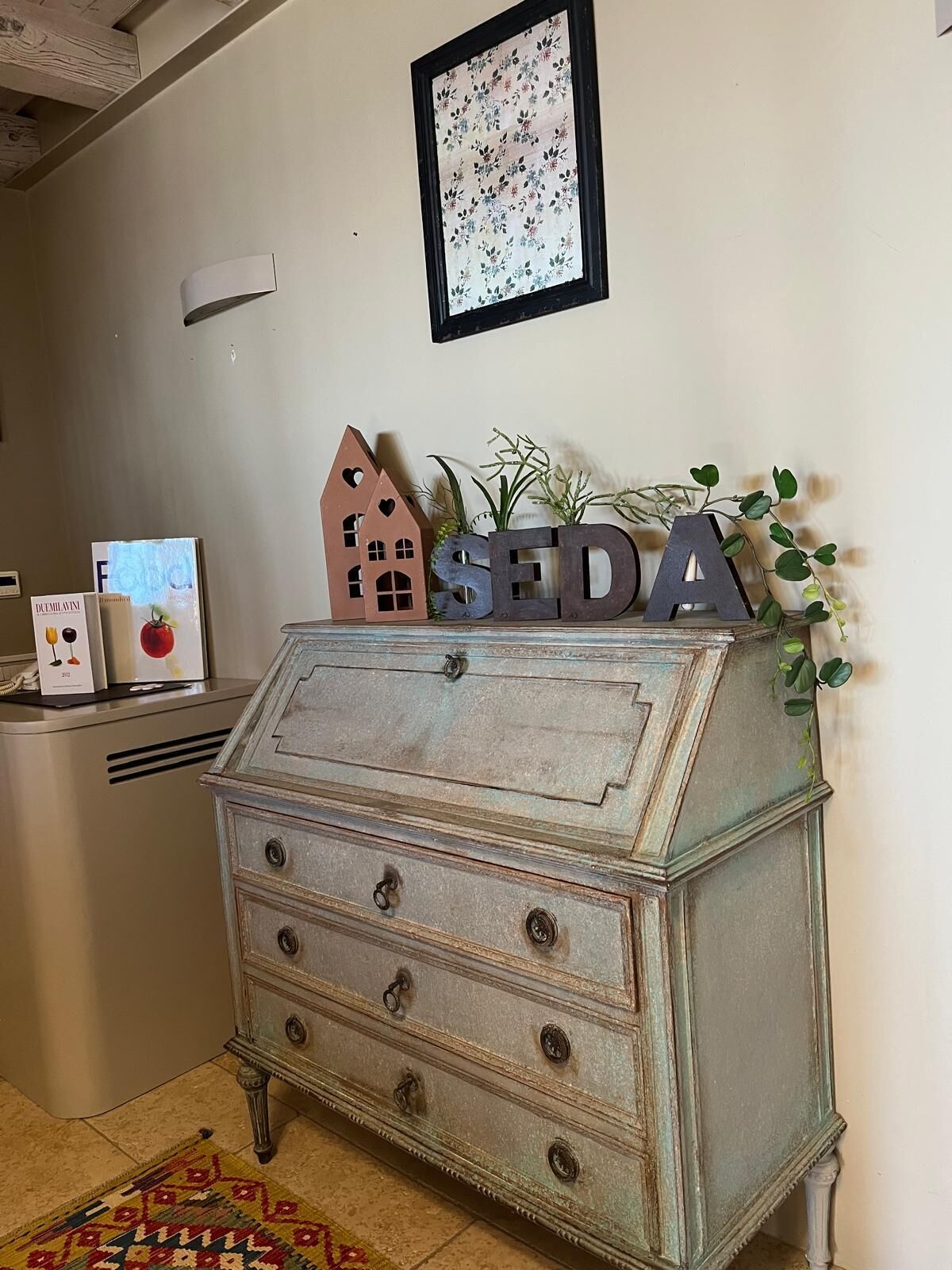 A vintage-style chest of drawers with the letters "SEDA" on top, a framed floral artwork above, and a small house-shaped decoration beside it.