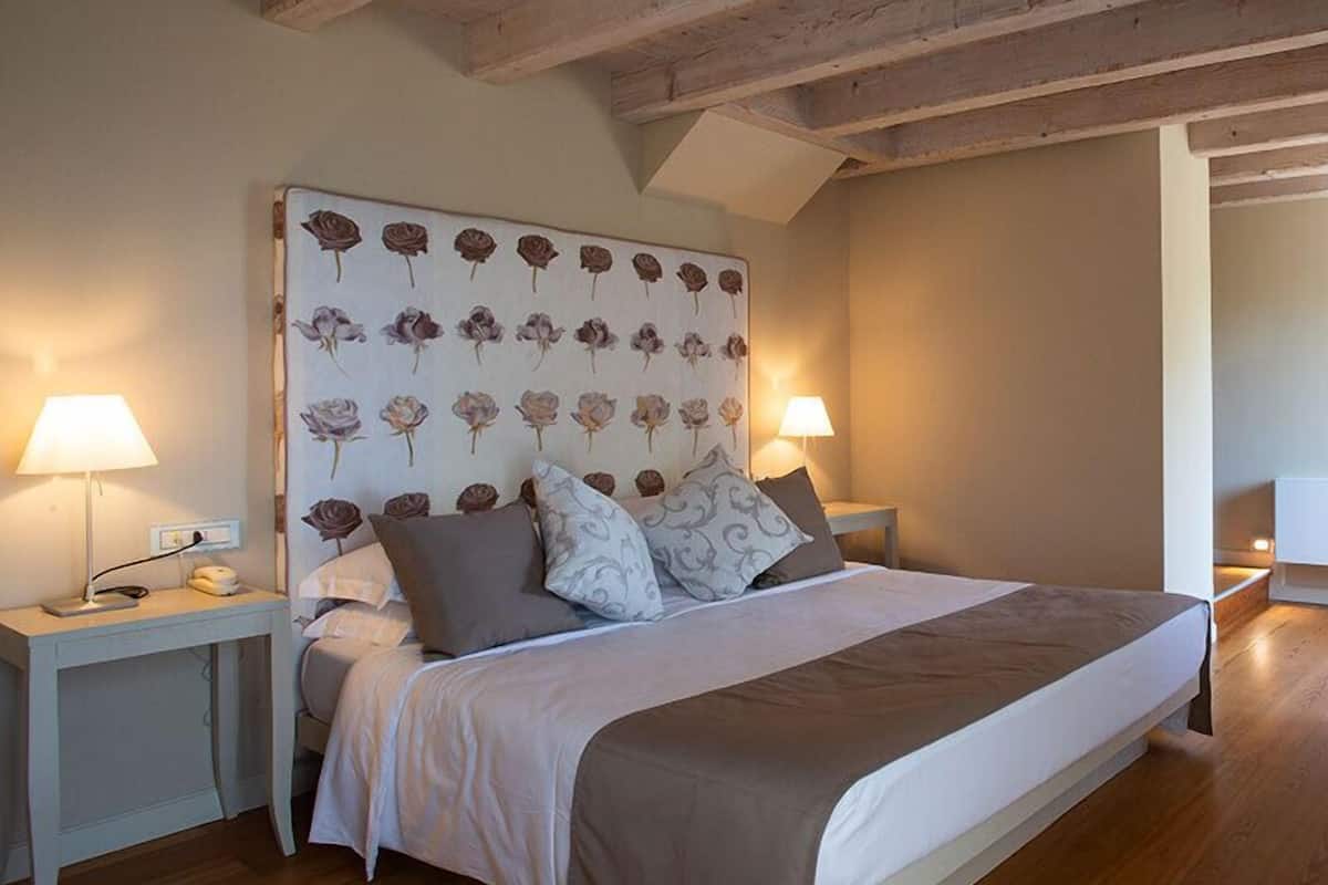 Een slaapkamer met een groot bed, houten balken in het plafond en een hoofdbord met bloemenprint. Er zijn twee nachtkastjes met lampen en een nachtkastje.