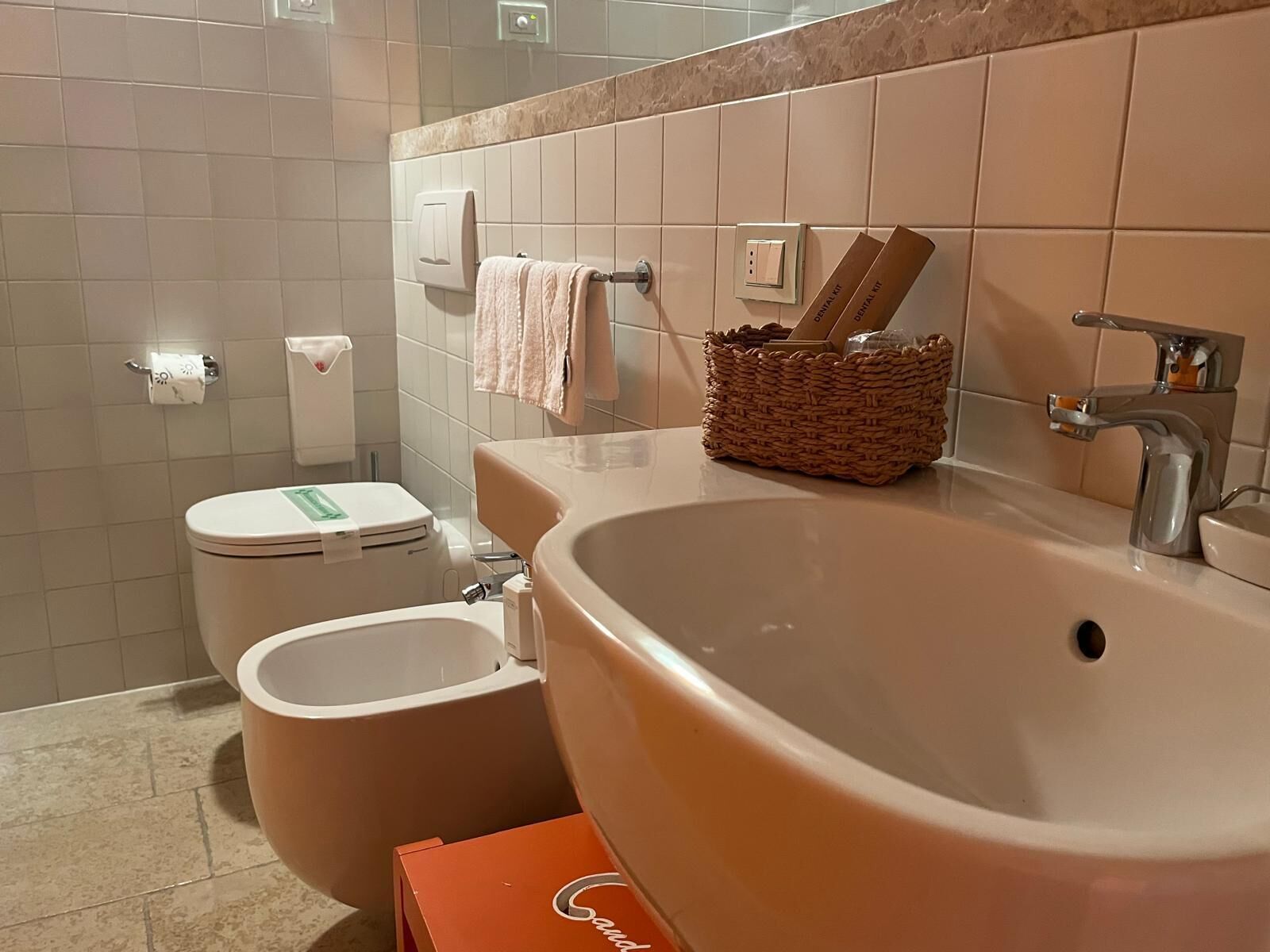 A bathroom with a large sink, a toilet, and a bidet. There is a towel hanging on the wall and a wicker basket with toiletries.