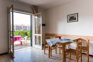 A room with a wooden table and chairs, a window with a view of buildings, and a door leading outside. - Cozy villa with private fenced garden and pool (Bibione)