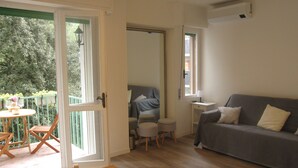 Una habitación con sofá gris, una mesita con una silla, un balcón con mesa y sillas, y una puerta que da a otra habitación.