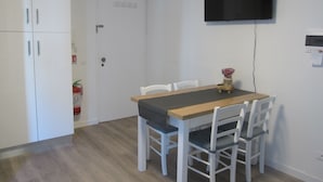 Una cocina pequeña y moderna con armarios blancos, una mesa de madera y sillas blancas. Un televisor de pantalla plana está montado en la pared sobre la mesa.