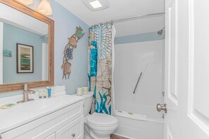 Une salle de bain avec un meuble-lavabo blanc, un grand miroir, un tableau encadré, une douche avec un rideau à motifs bleus et blancs, et des toilettes.