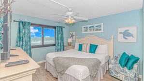 Un dormitorio con cama, ventilador de techo, ventana con cortinas, mesita de noche, lámpara y un cuadro enmarcado de una tortuga.