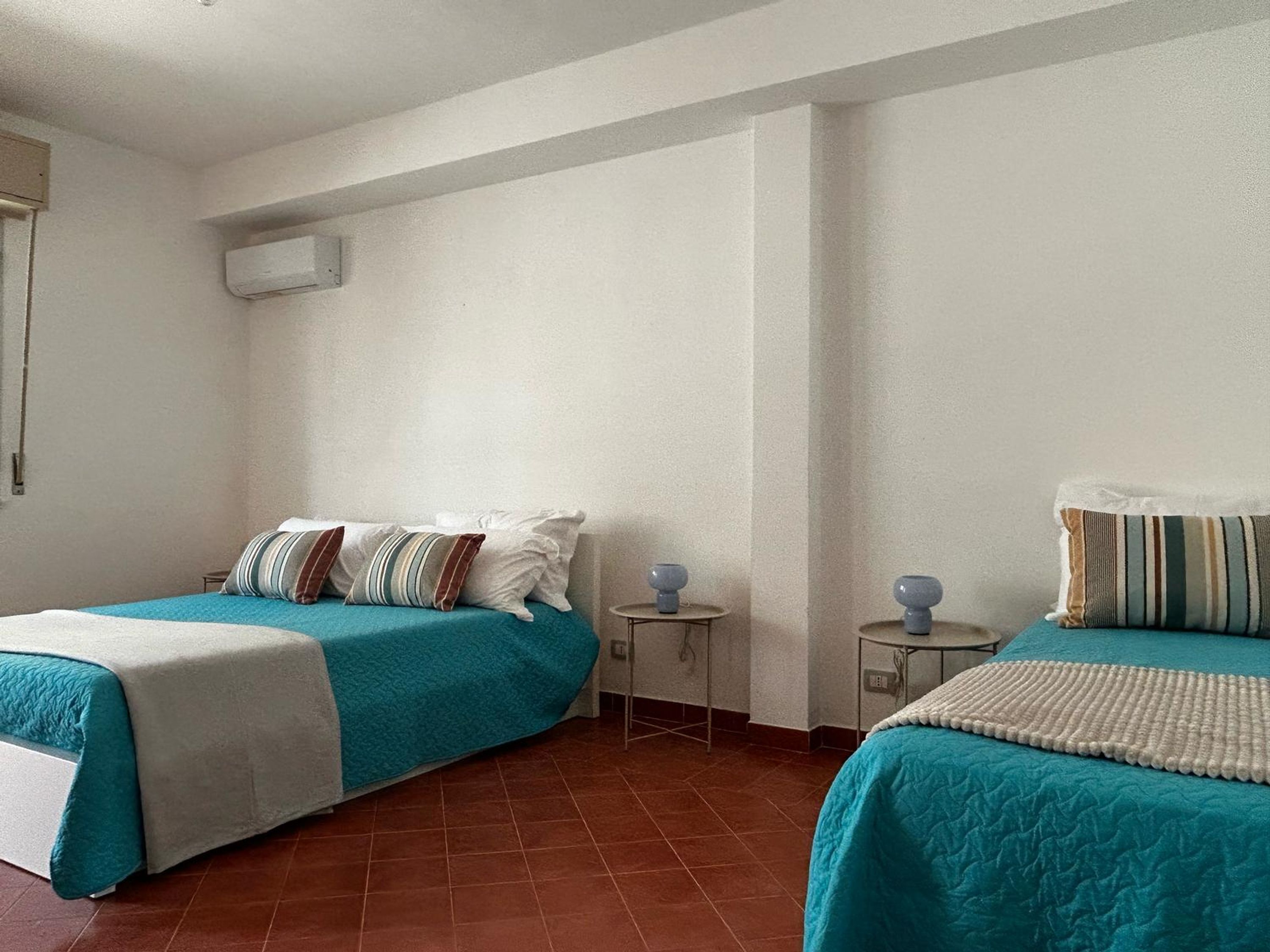 Une chambre d’hôtel avec deux lits, chacun recouvert d’une couvre-lit turquoise et agrémenté d’oreillers blancs, une petite table ronde avec une lampe, et un climatiseur fixé au mur.