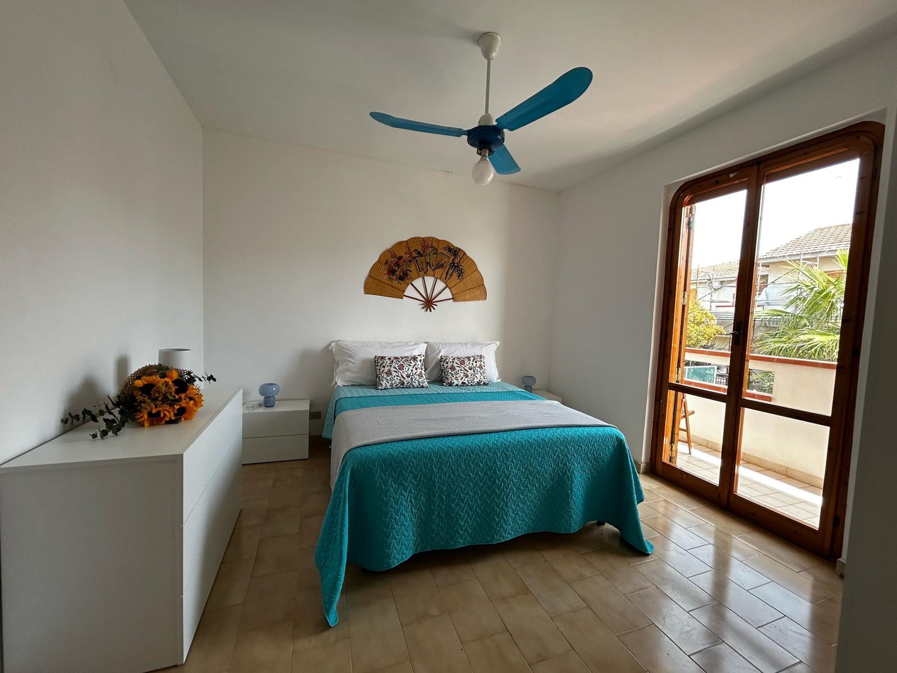 Een slaapkamer met een bed, een plafondventilator, een raam en een ventilator aan de muur.