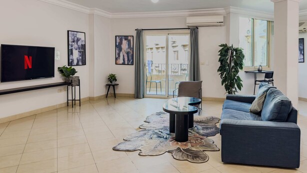 A living room with a flat-screen TV, a sofa, a coffee table, and a large window with a view of the outside. - Xuru Stays I Heliopolis Rock (Cairo)