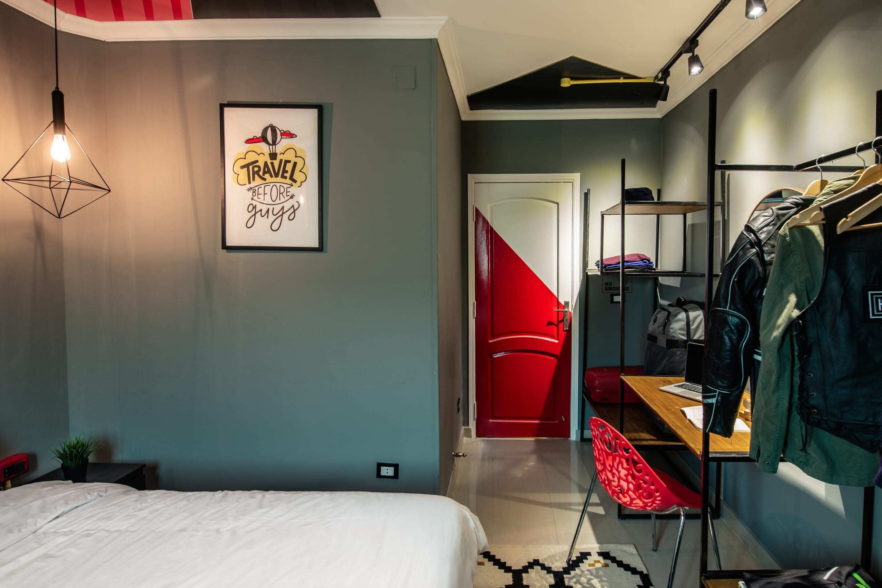 A bedroom with a bed, a red chair, a desk with a laptop, a clothing rack, and a red door.