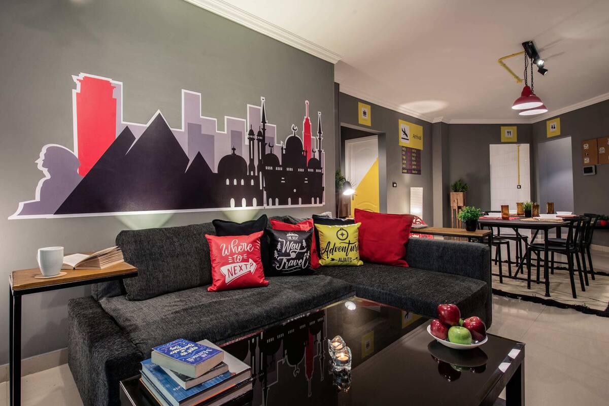 Een moderne woonkamer met een grijze bank, een salontafel met boeken en een glas, en een stadsgezicht muurschildering op de muur.