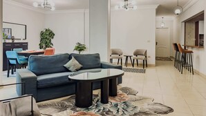 A modern living room with a blue sofa, a glass coffee table, and a dining area with blue chairs. There are two pendant lights, a large plant, and a view into another room. - Xuru Stays I Heliopolis Rock (Cairo)