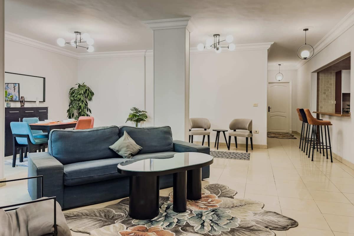 Een moderne woonkamer met een blauwe bank, een glazen salontafel en een eethoek met blauwe stoelen. Twee hanglampen, een grote plant en zicht op een andere kamer.