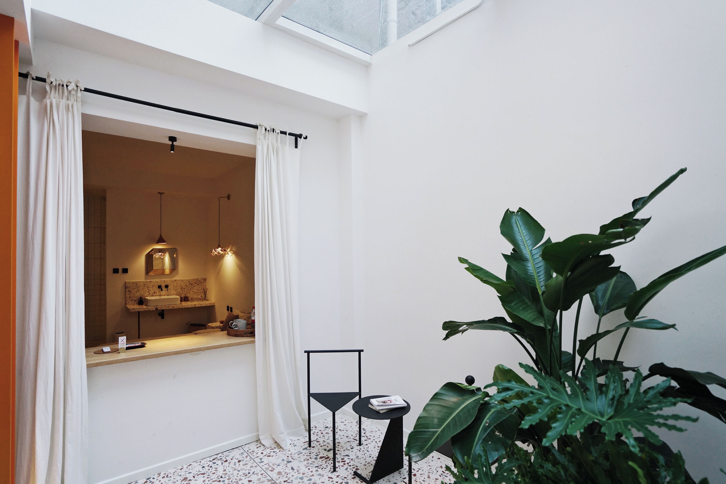 Un baño moderno con tragaluz, cortinas blancas, una mesita con un libro y una planta grande.