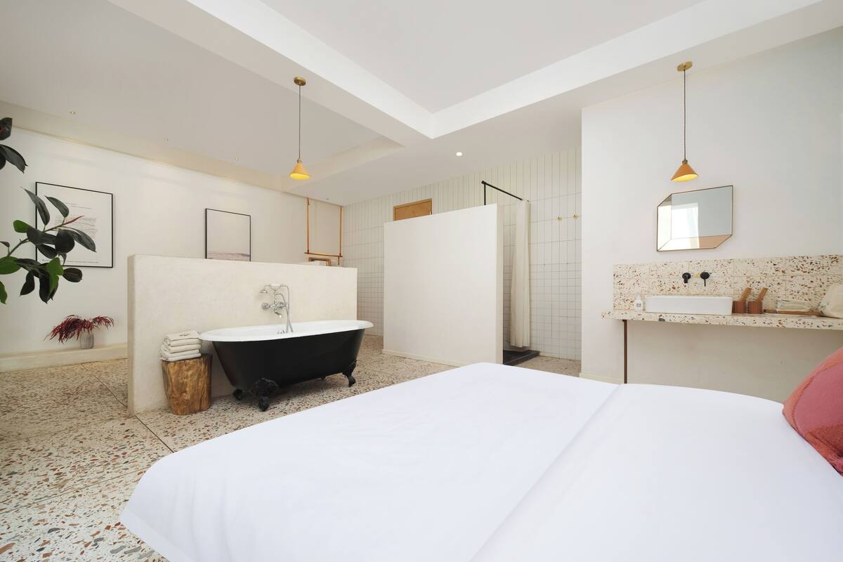 A modern bedroom with a large bed, a black clawfoot bathtub, a white vanity with a mirror, and a ceiling with recessed lighting.