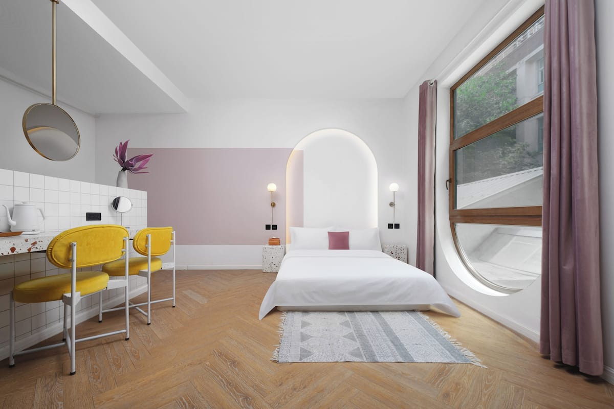 A modern bedroom with a bed, a desk, chairs, a mirror, and a window with curtains.