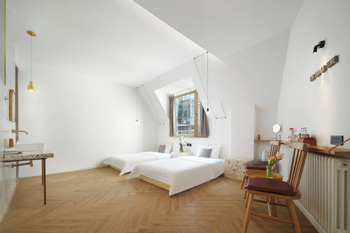 A modern, minimalist bedroom with two beds, a wooden floor, a skylight, and a small table with a chair.