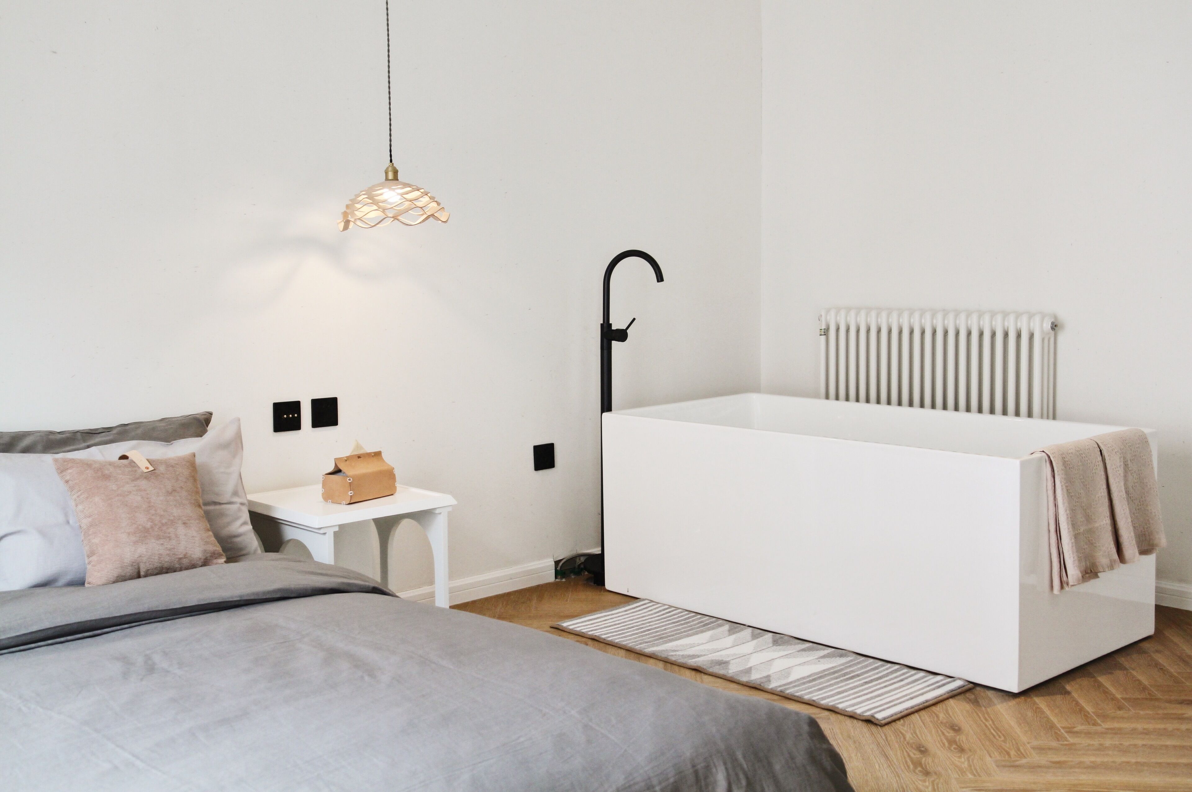 A modern bedroom with a white bed, a bedside table, a hanging light fixture, and a radiator.