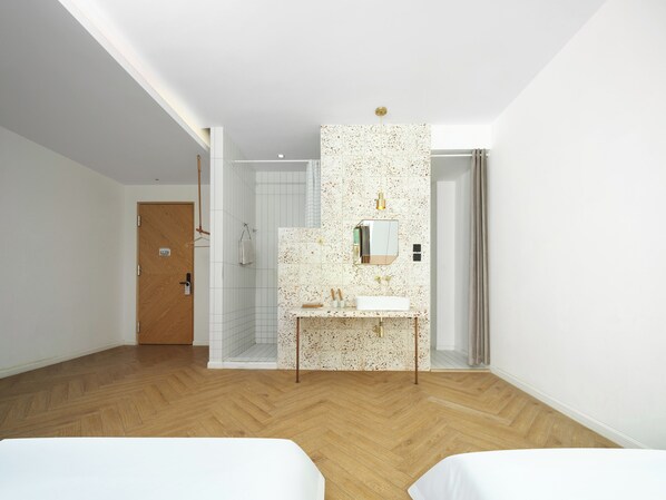 Una habitación de hotel moderna con suelo de madera, cama blanca, baño con ducha y una pequeña mesa con espejo y lavamanos.
