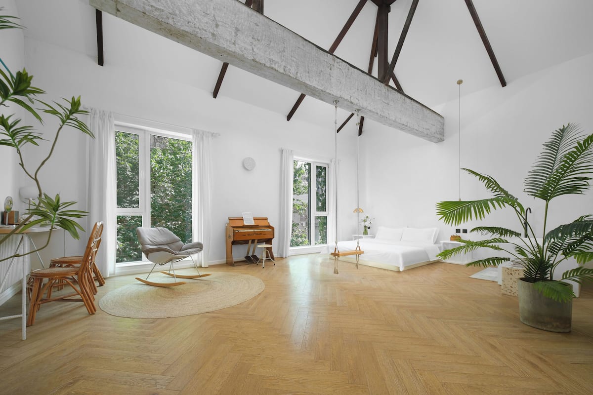A spacious room with large windows, a wooden floor, a piano, a sofa, and several potted plants.