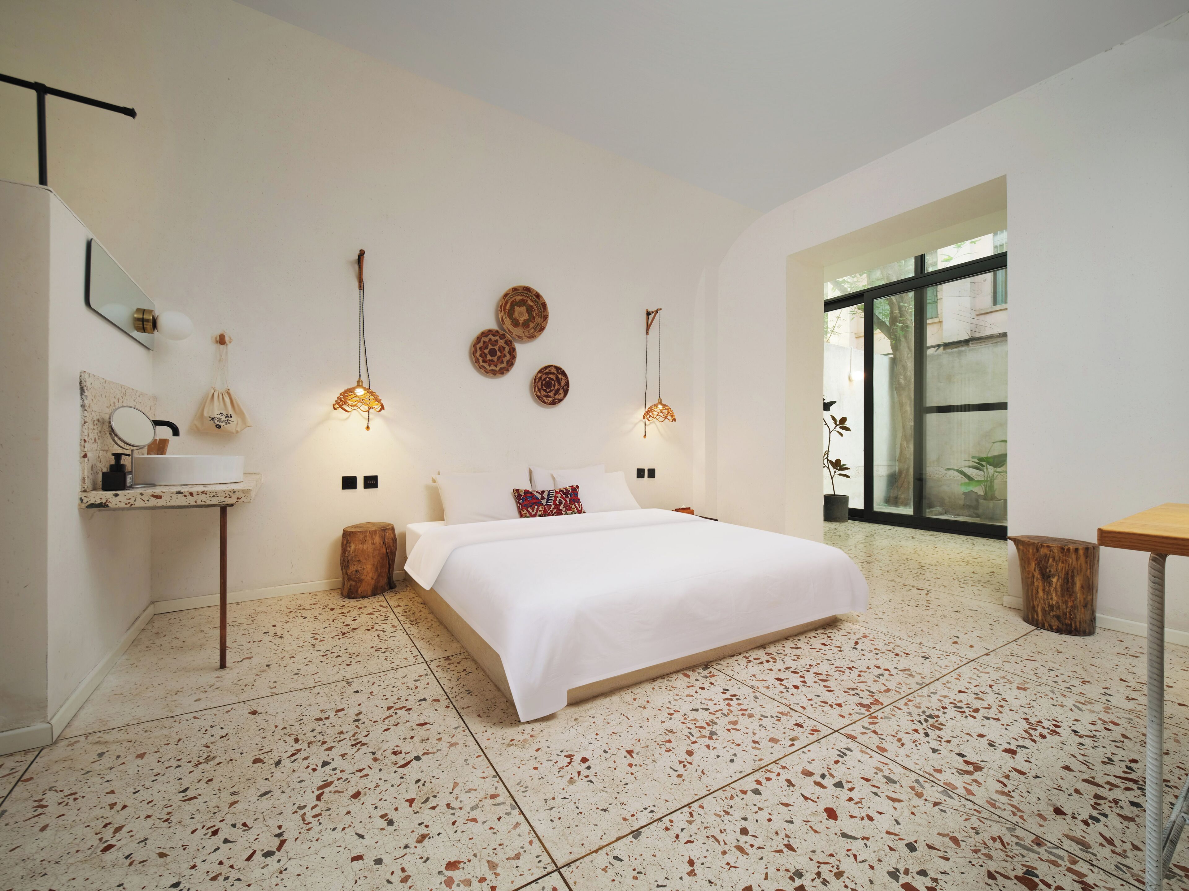 A modern bedroom with a large bed, a bedside table, a mirror, and a small desk. There are two wall-mounted lamps and a large window with a view of the outside.