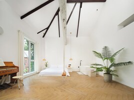 A spacious bedroom with a wooden floor, a piano, a bed, a hanging light fixture, and a large potted plant.