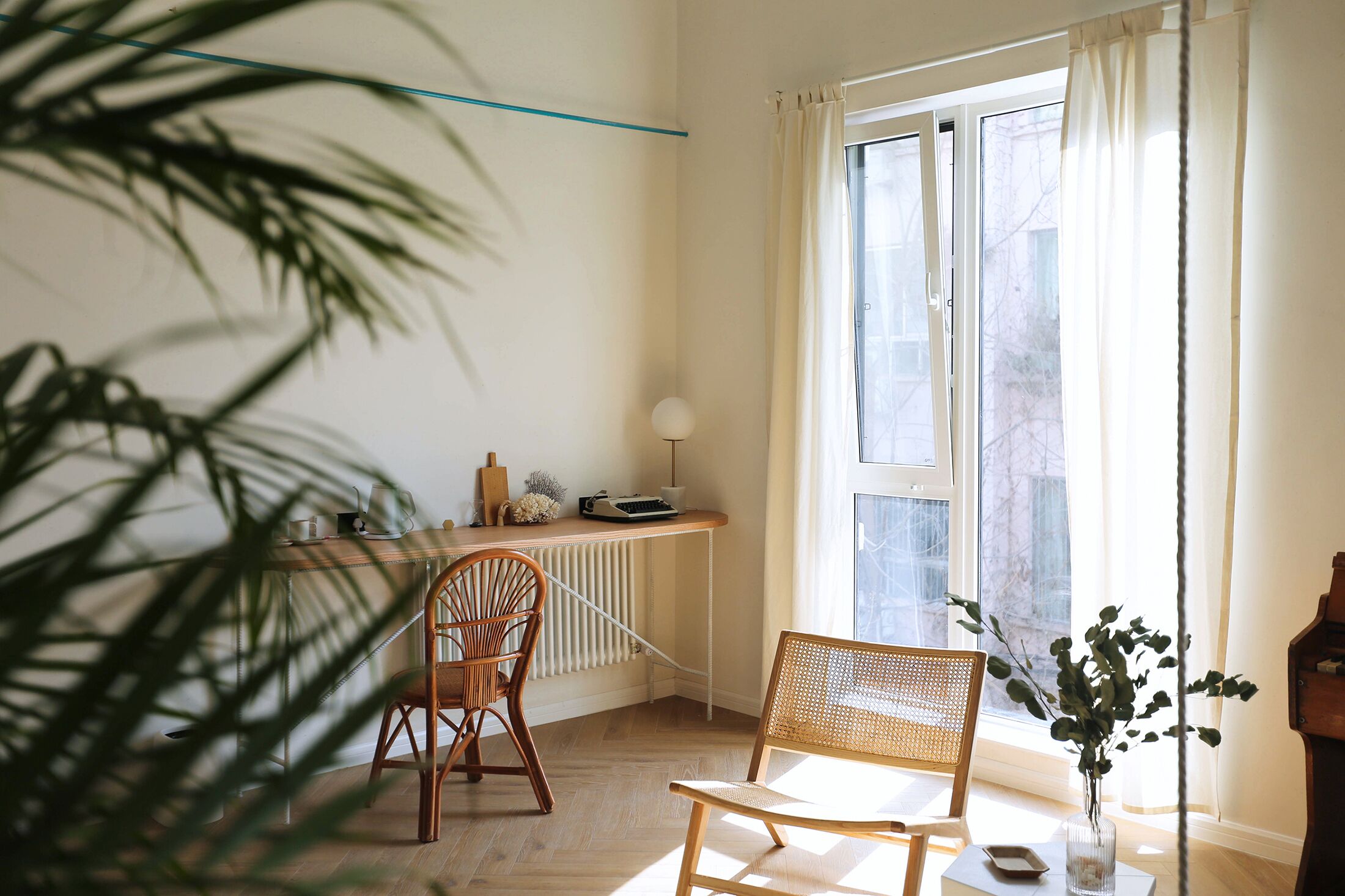 Una habitación con piso de madera, un radiador, un escritorio con lámpara, dos sillas de mimbre y una ventana con cortinas.