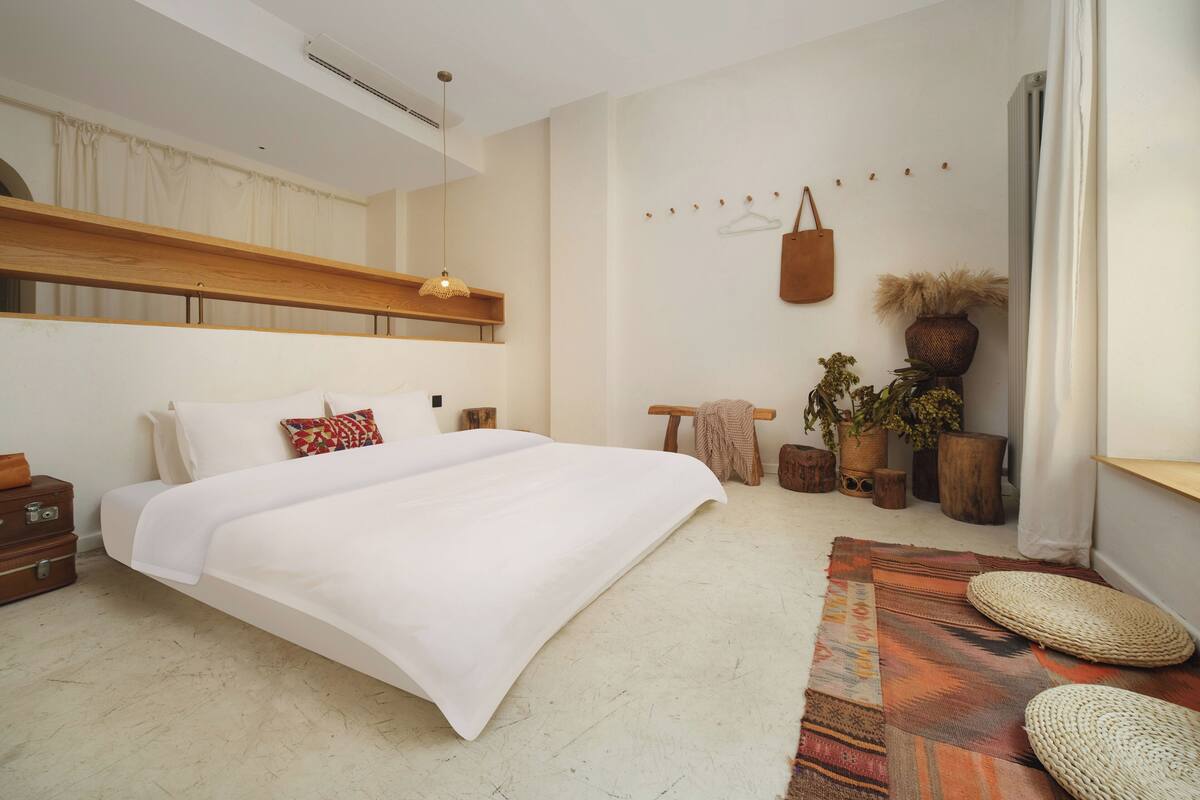 A modern bedroom with a large bed, a wooden headboard, a hanging light fixture, a bedside table, a small wooden stool, and a potted plant.