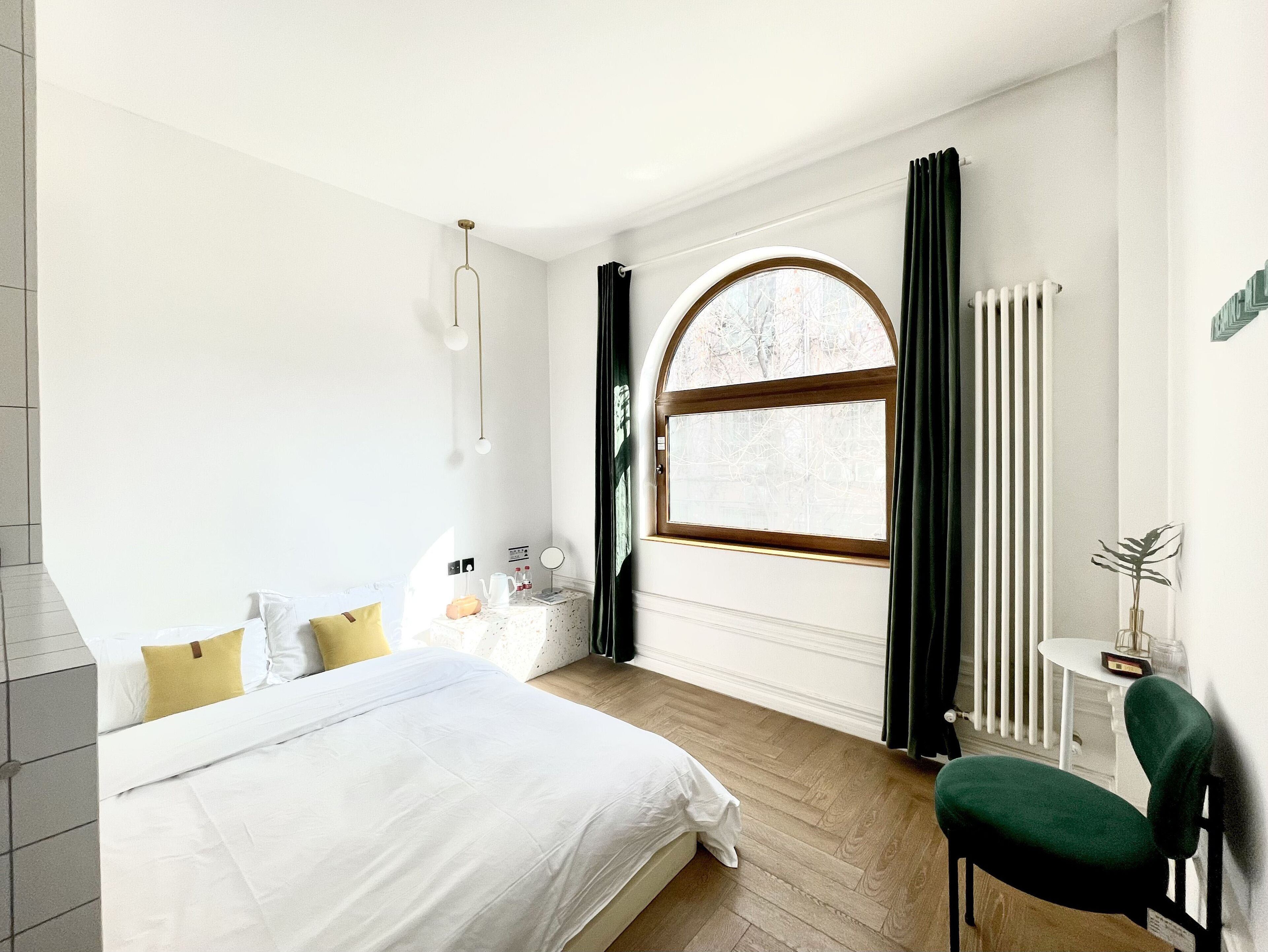 Une chambre à coucher avec un grand lit, une table de chevet avec une lampe, une fenêtre avec des rideaux, un radiateur et une chaise.