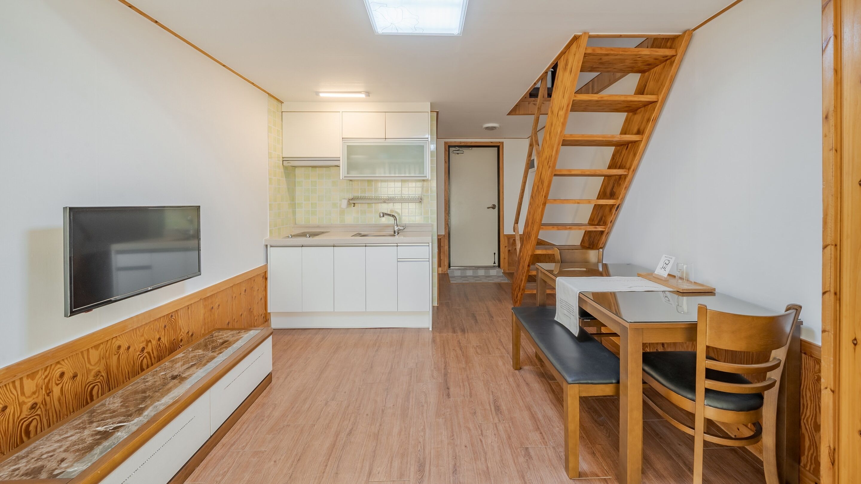 A compact kitchen with a built-in TV, a dining area with a bench and chairs, and a wooden staircase.