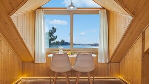 A room with a wooden interior, a table with two chairs, a window with curtains, and a view of a body of water and trees. - Yeongheungdo Rainbow Ocean View Pension (Incheon)