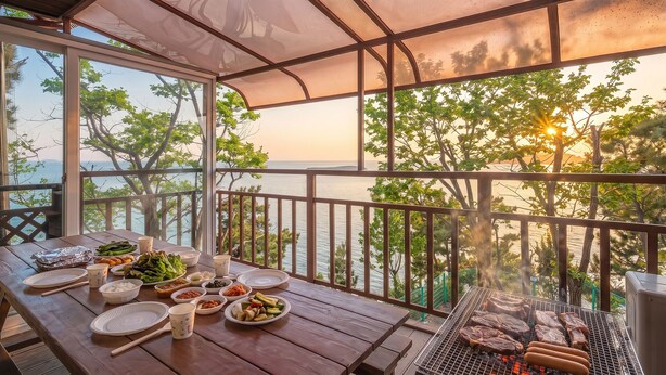 An outdoor dining area with a view of the sea and trees, a grill with meat, and various dishes on the table. - Yeongheungdo Rainbow Ocean View Pension (Incheon)