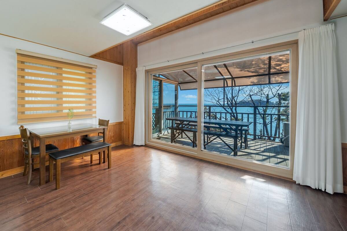 A room with a wooden table and chairs, a window with blinds, and a large sliding glass door leading to an outdoor deck with a view of the sea.