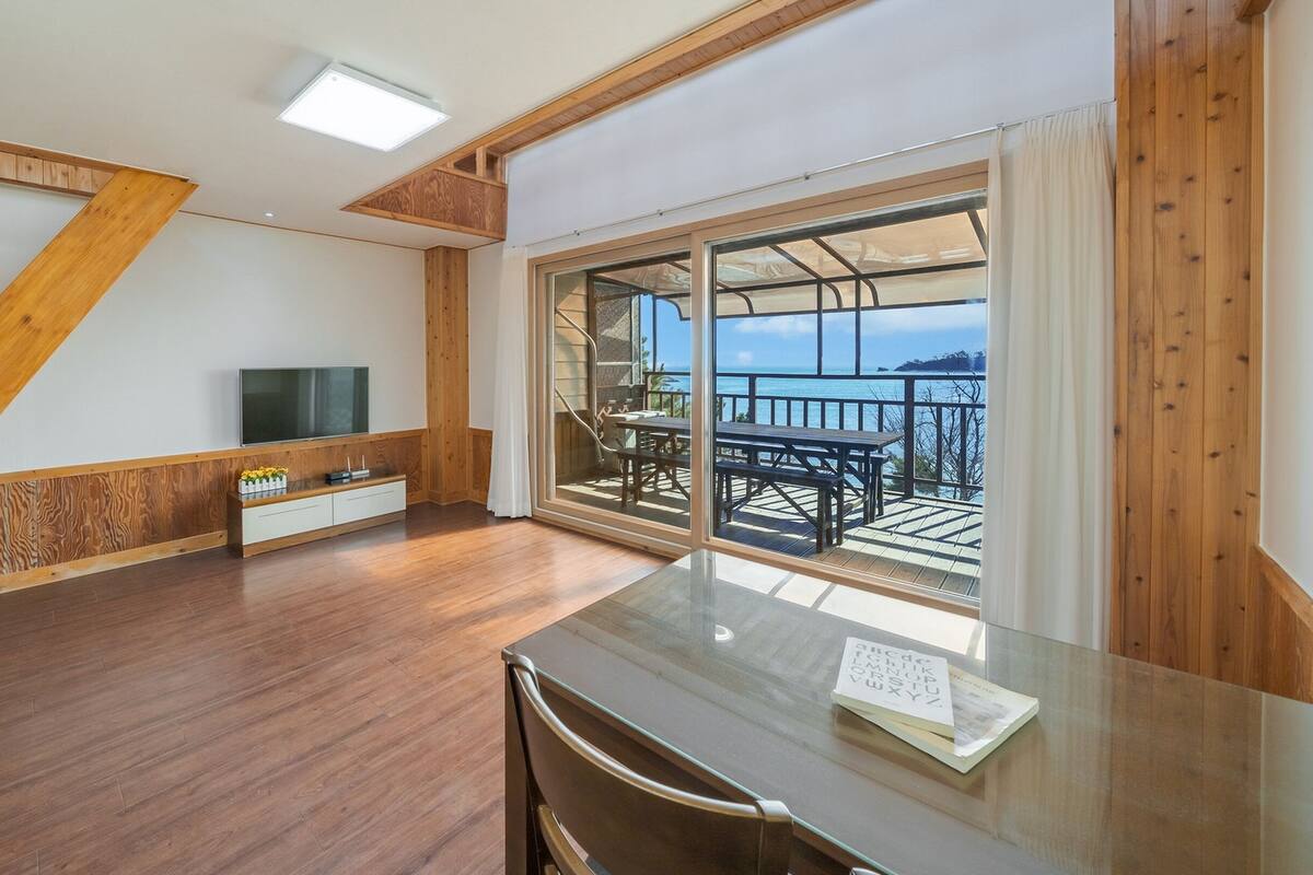 A modern room with a wooden floor, a flat-screen TV, a glass table, and a balcony with a view of the sea.