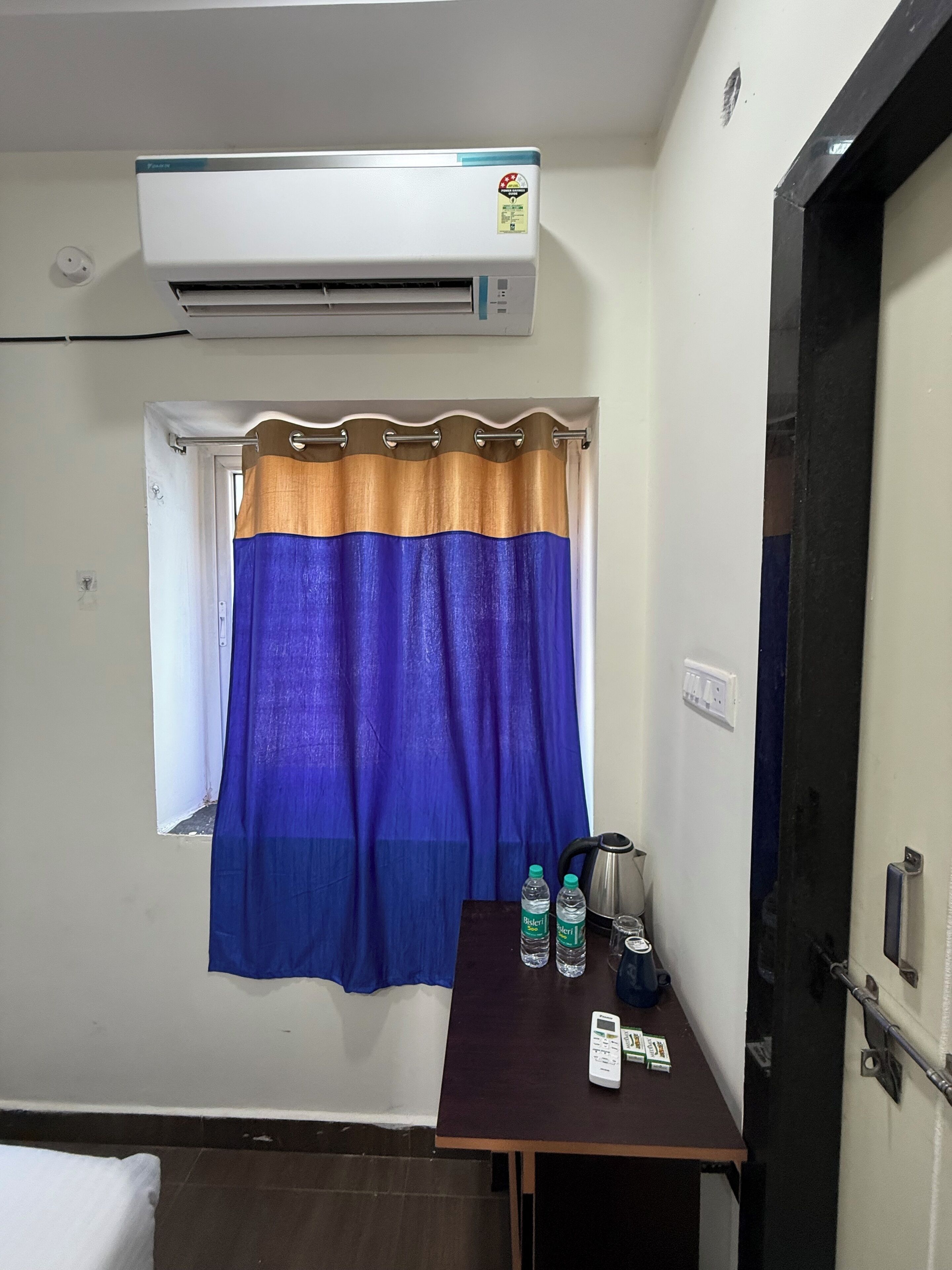A hotel room with a window, an air conditioner, a curtain, a table with a kettle, bottles, and a remote control.