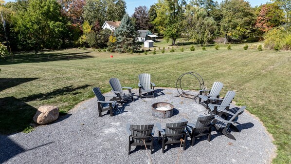 A circular seating area with Adirondack chairs around a fire pit, set on a gravel surface with a grassy lawn and trees in the background.