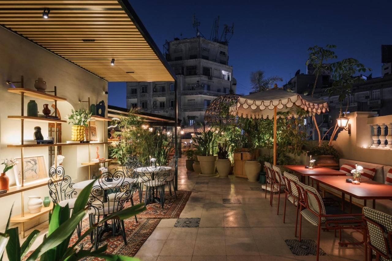 Un área de comedor al aire libre con mesas y sillas, rodeada de plantas en macetas y una pérgola de madera.