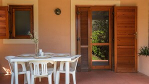A patio with a table and chairs, a window with shutters, and a door with a view of greenery.