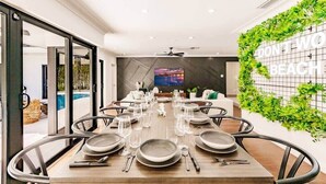 Un espace de repas moderne avec une longue table dressée pour un repas, un téléviseur en arrière-plan et un mur orné d’une pancarte indiquant : « N’AYEZ PAS D’INQUIÉTUDE, VOUS ÊTES À LA PLAGE ».