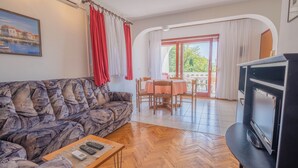 A living room with a patterned sofa, a wooden coffee table with remotes, a TV, and a dining area with a table and chairs. - Apartment Miskic 1 (Jadranovo)
