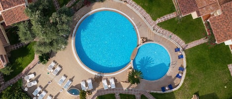 An aerial view of a resort with two large swimming pools, surrounded by lounge chairs and a well-maintained garden.