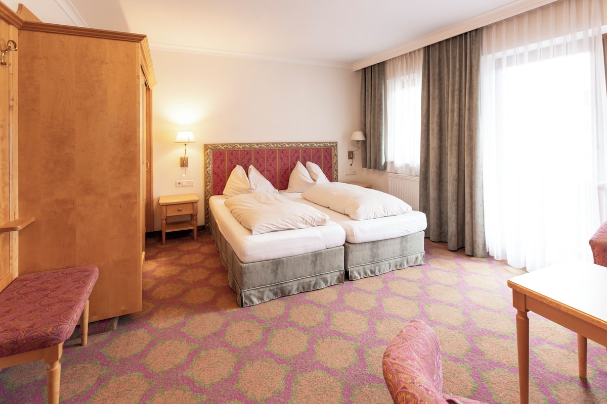 Habitació d'hotel amb llit doble, un armari de fusta, una petita taula de nit amb una làmpada, una cadira i una taula.