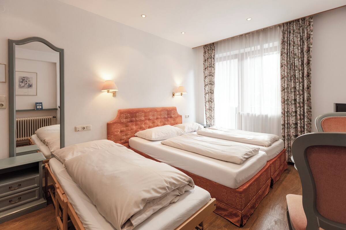Habitació d'hotel amb dos llits individuals, una tauleta de nit de fusta, una cadira, un mirall i una finestra amb cortines.