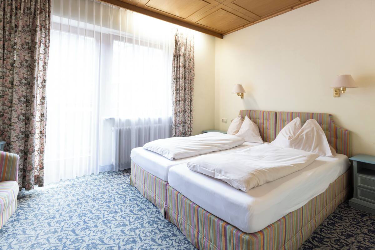 Habitació d'hotel amb un llit, dues taules de nit, una cadira i una finestra amb cortines.